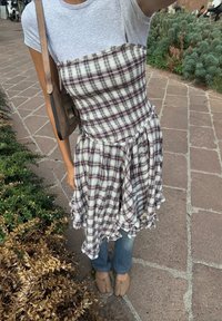 Geruite jurk in wit en bordeaux met een rimpelzoom, gedragen over een grijze t-shirt, gecombineerd met blauwe jeans en beige ballerina's. Schoudertas zichtbaar.
