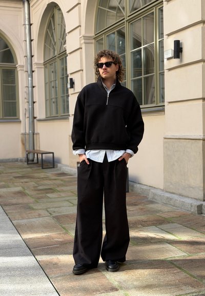 Hombre con cabello rizado y gafas de sol que lleva pantalones negros oversize y un suéter con cremallera sobre una camisa a rayas, de pie en un camino de piedra junto a un edificio.