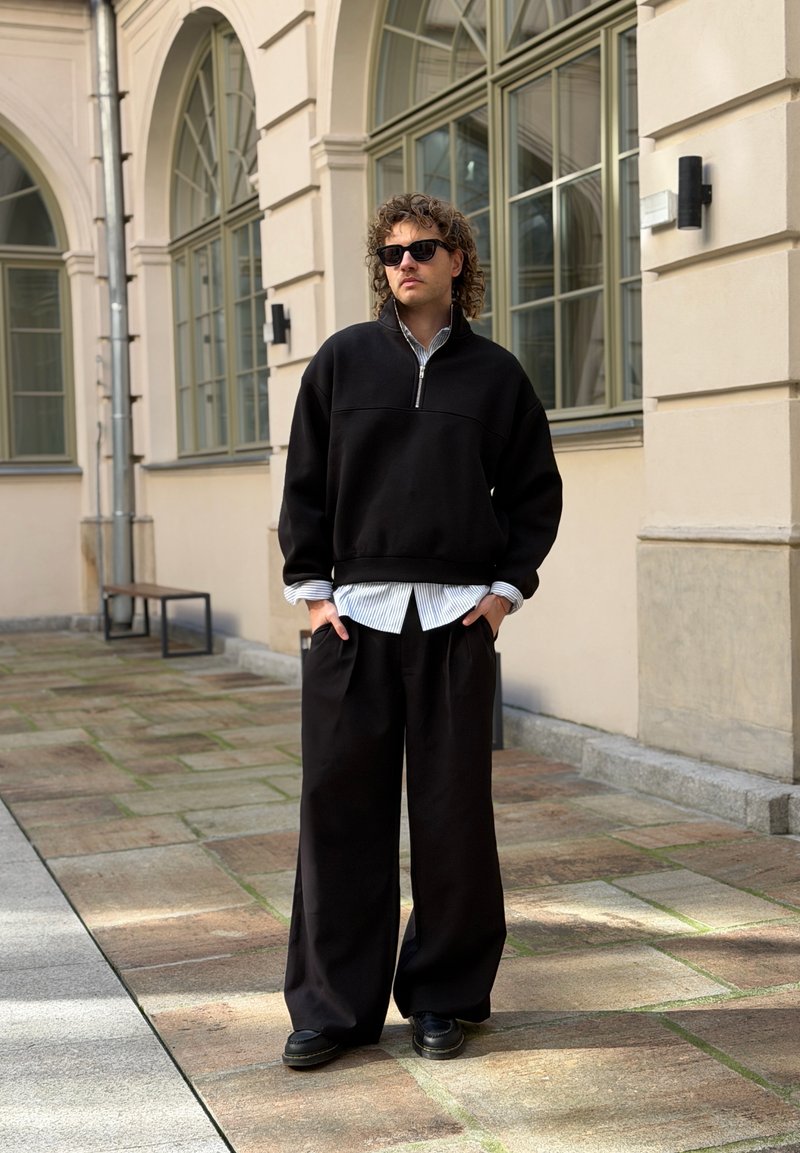 Man met krullend haar en zonnebril die zwarte oversized broek en een vest met rits draagt over een gestreept overhemd, staand op een stenen pad bij een gebouw.