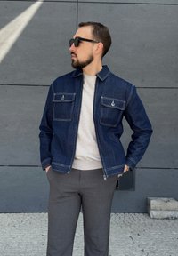 Chaqueta de denim azul oscuro con costuras en contraste, dos bolsillos en el pecho y cierre de cremallera, combinada con pantalones grises y un suéter ligero.