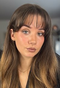 Portrait en gros plan avec de longs cheveux bruns ondulés. Présente une peau claire, des yeux verts et un anneau de septum. Porte de petites boucles d'oreilles en anneau et un collier en croix.