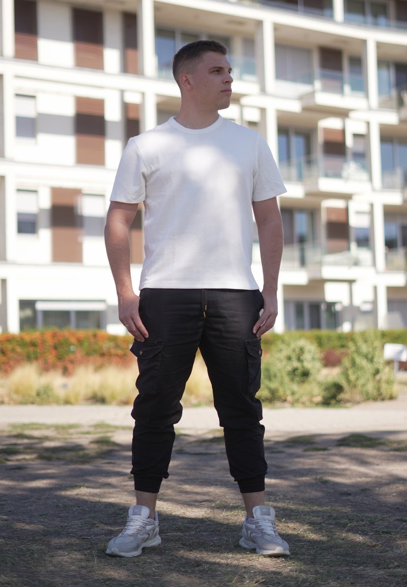 Jeune homme portant un t-shirt blanc, un pantalon cargo noir et des baskets blanches se tenant dehors devant un immeuble résidentiel.