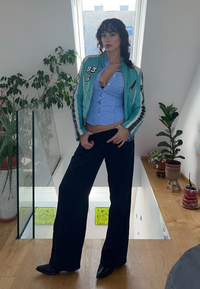 Una chaqueta de cuero turquesa con franjas racing sobre una camisa de rayas azules y blancas, combinada con pantalones de pierna ancha negros y zapatos negros.