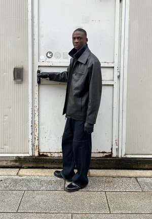 Chaqueta de cuero negra con un cuello amplio, complementada con unos vaqueros oscuros holgados y zapatos negros. Guantes cubren las manos; está de pie junto a una puerta de metal.