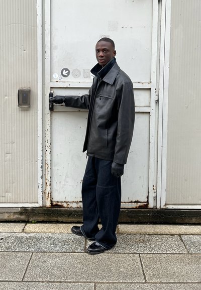Chaqueta de cuero negra con un cuello amplio, complementada con unos vaqueros oscuros holgados y zapatos negros. Guantes cubren las manos; está de pie junto a una puerta de metal.