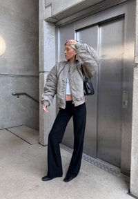 Grijze pufferjack met een rits, witte cropped top, zwarte wijde broek en zwarte schoenen, staande bij een lift in een betonnen omgeving.