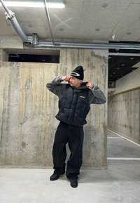 Gilet a vento nero con quattro tasche frontali sopra un maglione grigio, abbinato a jeans neri larghi e stivali neri in un contesto di cemento.