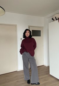 Pull à col roulé en tricot bordeaux, pantalon large gris et chaussures noires, posés dans une pièce aux couleurs neutres avec un sol en bois clair.