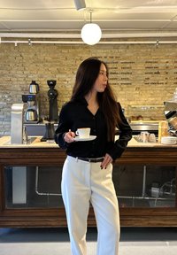 Schwarzes Hemd mit Knöpfen, weiße Hose, hält eine Tasse auf einer Untertasse. Im Hintergrund ist eine Holztheke und Kaffeemaschinen zu sehen. Hell beleuchteter Raum.