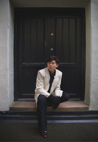 Hvit blazer over en svart skjorte, svarte bukser og hansker. Sittende på trappetrinn foran en svart tremur med sølvfarget beslag. Brune sko.