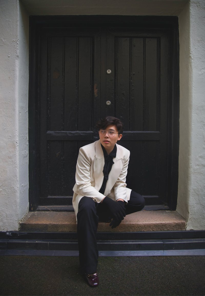 Bijeli sako preko crne košulje, crne hlače i rukavice. Sjedi na stepenicama ispred crnih drvenih vrata sa srebrnim okovima. Smeđe cipele.