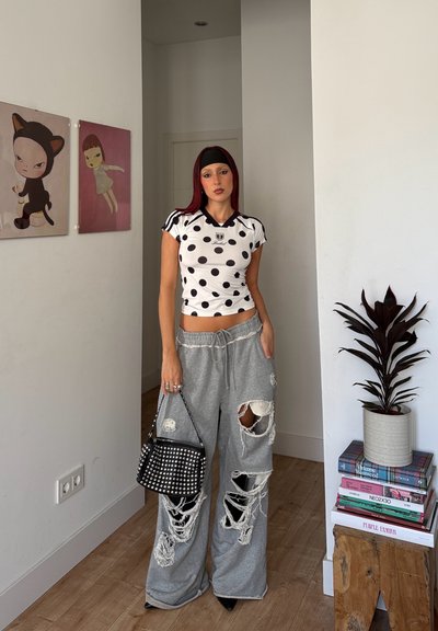 Mujer con pelo rojo que lleva una blusa de lunares, pantalones de pierna ancha grises rasgados, diadema negra, sosteniendo un bolso negro con tachuelas en el interior.