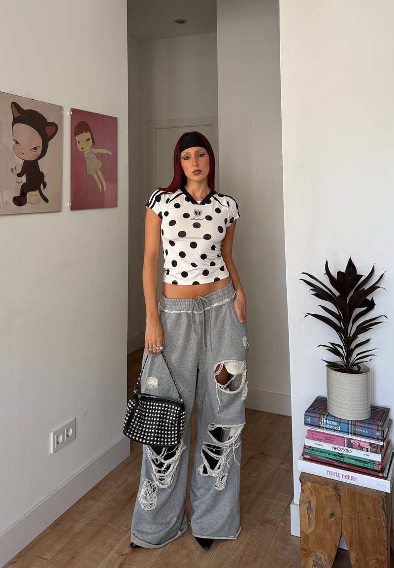 Punane juustega naine kannab täpilist särki, rebenditega halli laiade jalgadega pükse, musta peapaela ning hoiab musta narmastega käsipakki siseruumis.