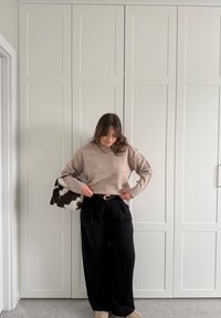 Femme ajustant un pull beige rentré dans un pantalon noir taille haute, tenant un sac à main noir et blanc à motif, debout devant des portes blanches à panneaux.