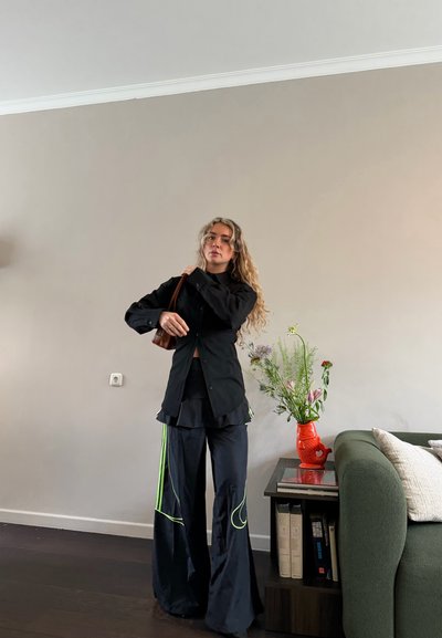 Mujer con cabello rubio largo, vistiendo camisa negra y pantalones amplios negros con rayas verdes, sosteniendo una bolsa marrón, de pie cerca de un sofá verde y una mesa auxiliar con un jarrón.