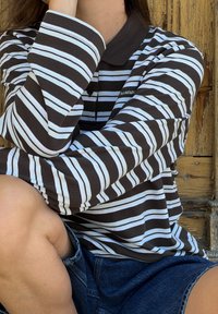 Pessoa sentada com um joelho elevado, vestindo uma polo de mangas longas preta e branca com riscas e calções de ganga, contra um fundo de madeira.