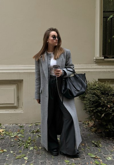 Abrigo largo de lana gris, blusa blanca, pantalones de pierna ancha negros, zapatos de plataforma negros, gafas de sol redondas, sosteniendo una taza de café, con un bolso negro.