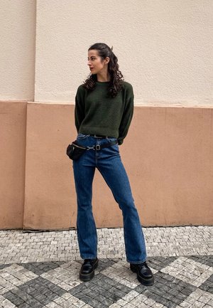 Suéter de punto verde, jeans acampanados azules con un cinturón, bolsa de cintura negra, botas de plataforma negras; de pie contra una pared texturizada y pavimento.