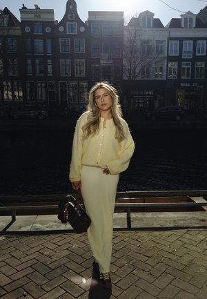 Jasnożółty wełniany kardigan, fakturowana biała spódnica, brązowe buty z żółtymi sznurówkami, trzymając wzorzysty torebkę, stojąc na ceglanej nawierzchni przy kanale.