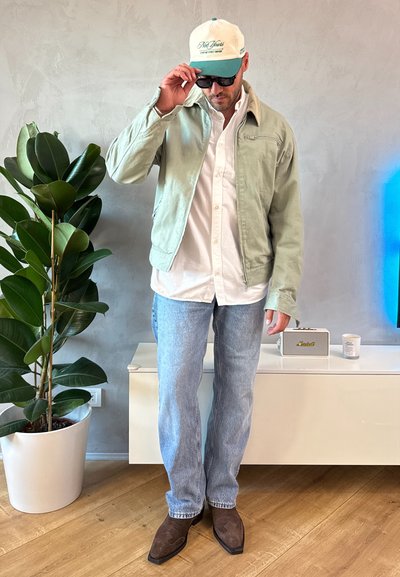 Chaqueta verde claro, camisa blanca, vaqueros azules, zapatos marrones y una gorra bicolor. Ambiente interior con plantas y decoración minimalista.