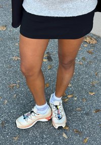 Witte en beige sportschoenen met zwarte veters en kleurrijke accenten. Draagt witte sportsokken en een zwarte rok, staand op een gestructureerd voetpad.