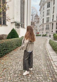 Veste beige surdimensionnée, chemise bleu clair, pantalons gris à jambes larges et baskets blanches. Chemin pavé et bâtiments dans un cadre urbain.