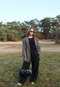 Giacca di lana marrone sopra una maglietta nera, pantaloni neri larghi, con una borsa a tracolla in pelle nera. Occhiali da sole e un ambiente naturale.