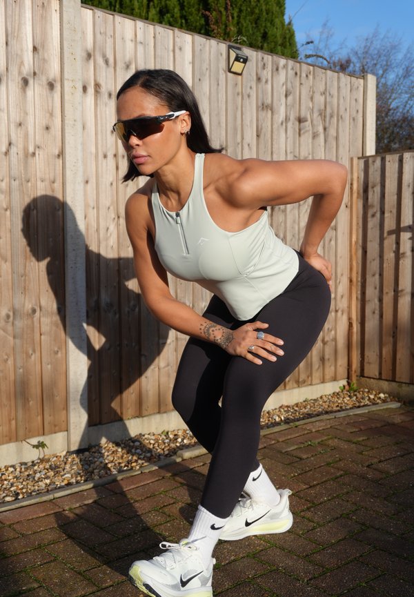 Mujer con ropa deportiva y gafas de sol estirándose al aire libre, inclinada hacia adelante con las manos sobre las rodillas, cerca de una cerca de madera en un día soleado.