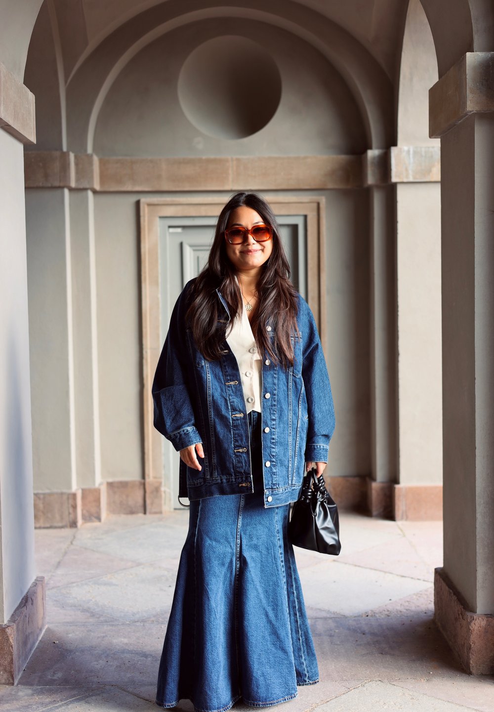 Denim jacket with a slightly oversized fit, paired with a long denim skirt. Black handbag and sunglasses. Grey architectural background.