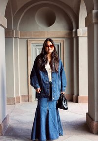 Denim jacket with a slightly oversized fit, paired with a long denim skirt. Black handbag and sunglasses. Grey architectural background.