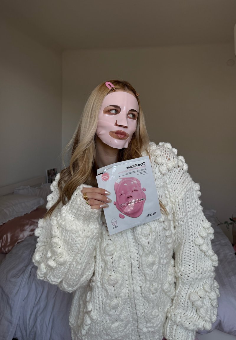 Femme aux longs cheveux blonds portant un pull blanc en maille épaisse, avec un masque facial rose, tenant son emballage dans une chambre.