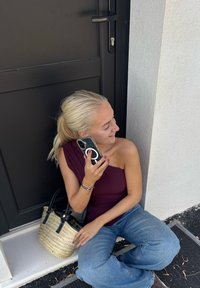 Kobieta w topie na jedno ramię w kolorze burgundowym siedzi przy czarnych drzwiach, trzymając telefon. Ma blond włosy i nosi niebieskie dżinsy z szerokimi nogawkami oraz słomkową torbę.