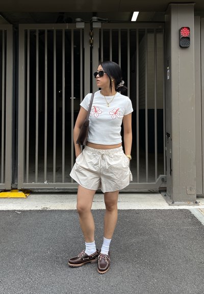 Camiseta blanca corta con estampados florales rojos, pantalones cortos beige con cintura elástica, calcetines blancos, zapatos marrones y un bolso marrón de hombro.