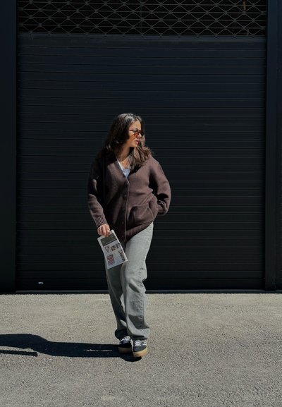 Cardigan de punto marrón sobre una blusa clara, pantalones de pierna ancha grises y zapatillas negras. Sosteniendo un periódico, de pie contra un fondo oscuro.