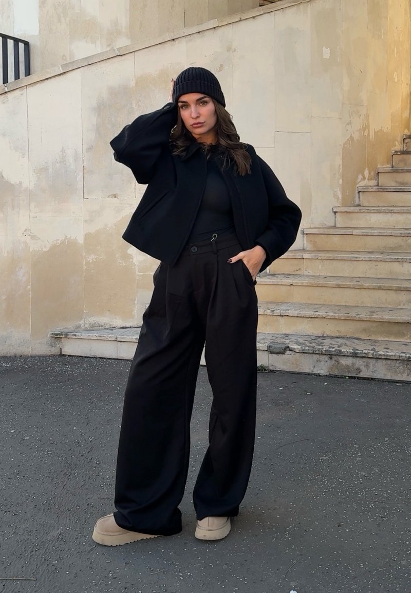 Donna in piedi all'aperto vicino a scalini in pietra consumati, indossa un berretto nero, una giacca nera oversize, pantaloni neri larghi e scarpe beige con piattaforma.