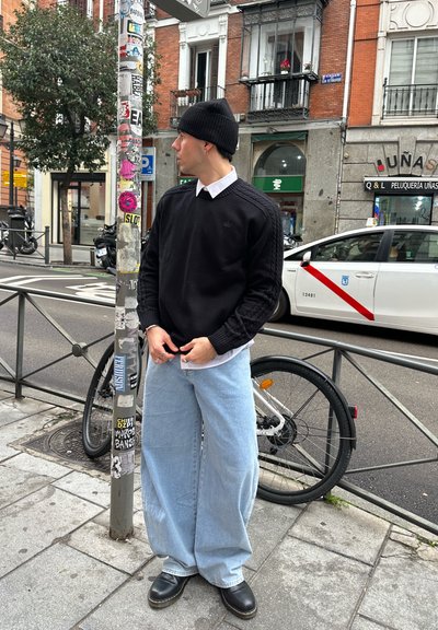 Joven con gorro negro y suéter, con pantalones anchos de mezclilla azul claro, de pie en la acera de la ciudad cerca de una bicicleta y un taxi.