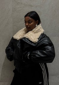 Sort læderjakke med en blød beige krave, oversized form og skinnende tekstur. Båret over sort tøj med hvide striber.