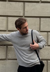 Grå strikket sweater med rund hals, struktureret mønster og ribbet kant. Sort skuldertaske, mørke bukser, foran en lys stenvæg.