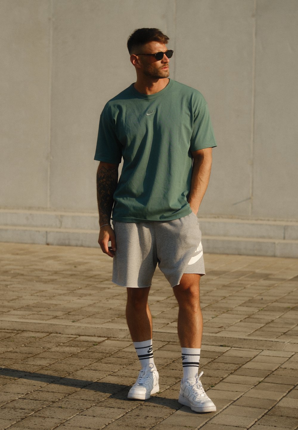 Zöld pamut póló, szürke Nike logós sportnadrág, fehér sneaker és fekete napszemüveg. Kényelmes sportos öltözet texturált járdán.