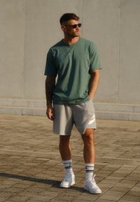 Zöld pamut póló, szürke Nike logós sportnadrág, fehér sneaker és fekete napszemüveg. Kényelmes sportos öltözet texturált járdán.