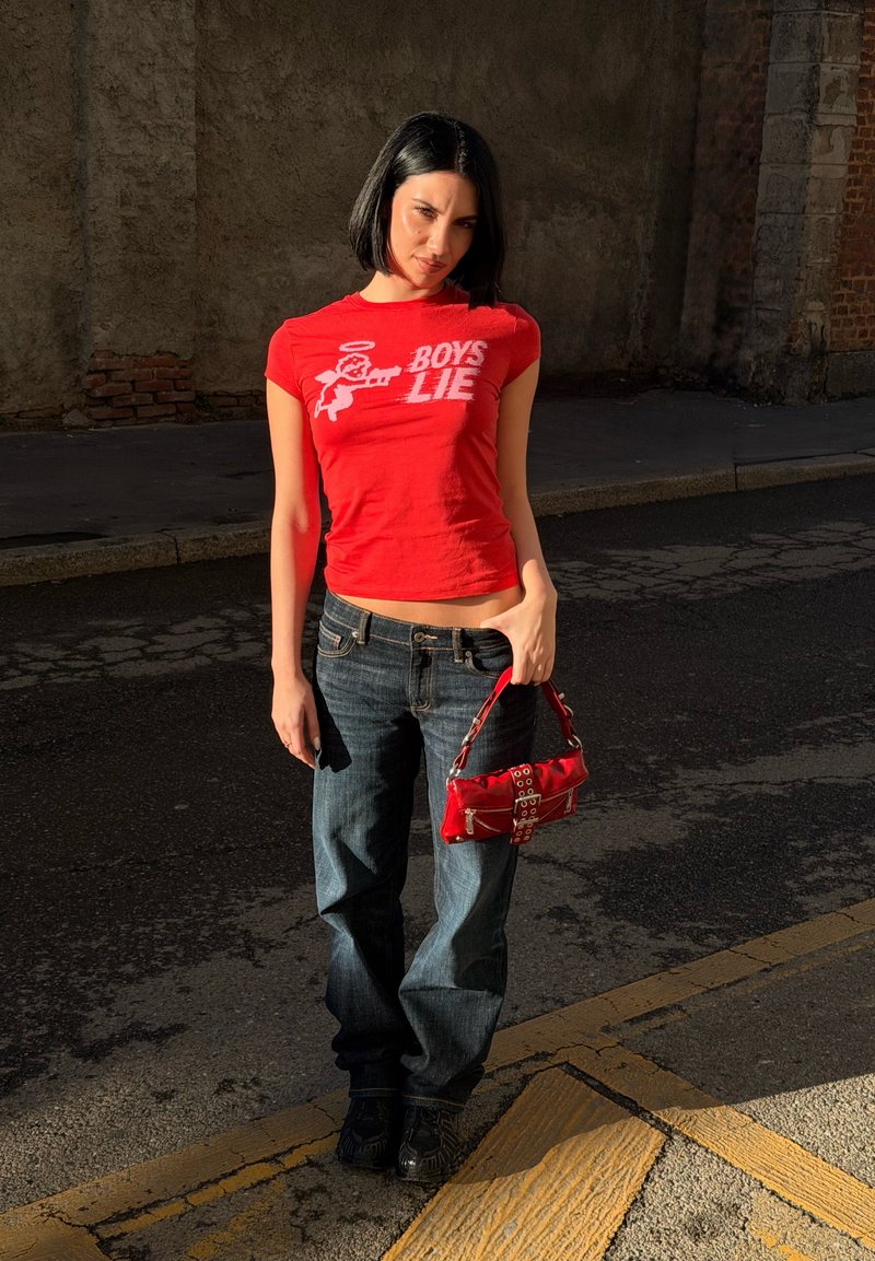 Νέα γυναίκα με κοντό μαύρο καρέ, φορώντας κόκκινο πουκάμισο με τη φράση «Τα αγόρια λένε ψέματα», μπλε τζιν, κρατώντας μια κόκκινη τσάντα, στέκεται σε έναν ηλιόλουστο δρόμο.