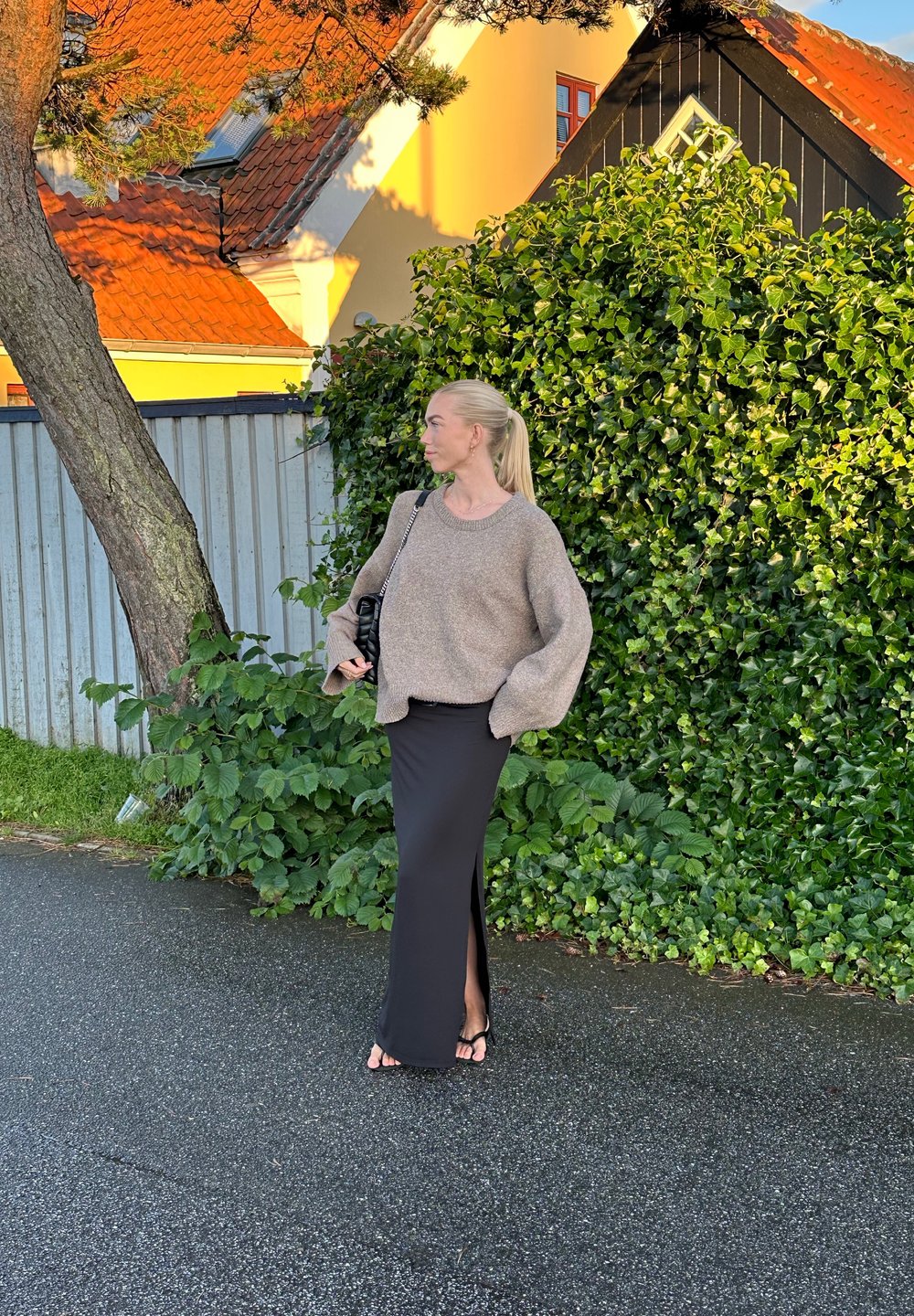 Hnědý oversize svetr a černá maxi sukně s bočním rozparkem. Černá taška s popruhem, ploché sandály. Zelené listoví a městské pozadí.