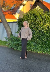 Hnědý oversize svetr a černá maxi sukně s bočním rozparkem. Černá taška s popruhem, ploché sandály. Zelené listoví a městské pozadí.