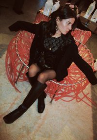 Κόκκινη, αφηρημένη καρέκλα από σύρμα με καμπυλωτό σχέδιο. Το μοντέλο φοράει ένα μαύρο τούλινο τοπ και μακριές, μαύρες μπότες, καθισμένο σε ένα υφασμάτινο χαλί.