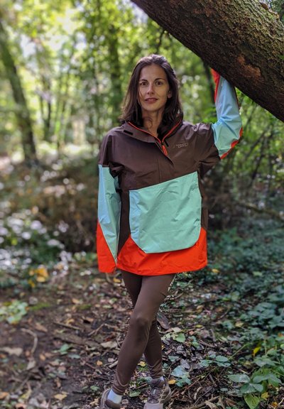 Mujer vestida con chaqueta y leggings marrones, turquesas y naranjas se encuentra en un camino del bosque, apoyando un brazo en una rama de árbol.
