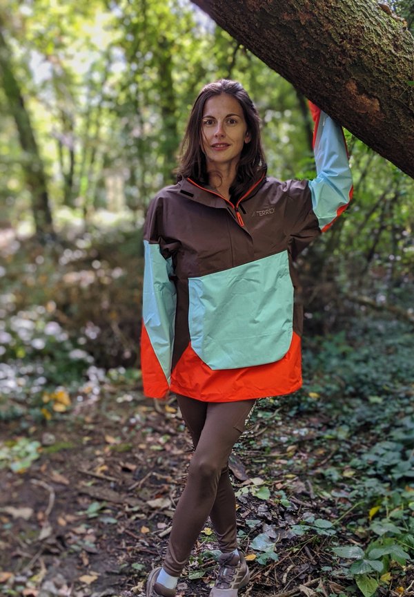 Chaqueta resistente al agua en marrón, azul claro y naranja; cuenta con media cremallera, un gran bolsillo frontal y puños ajustables. Llevada con leggings marrones.