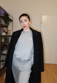 Sweat-shirt gris et pantalon de survêtement avec logo Adidas sous un long manteau noir. Design simple, texture lisse, cadre intérieur avec étagères de livres.