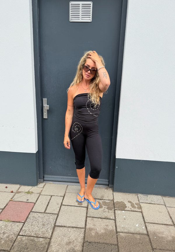 Femme aux longs cheveux blonds et lunettes de soleil, portant une combinaison noire sans bretelles et des tongs bleues, debout devant une porte gris foncé.