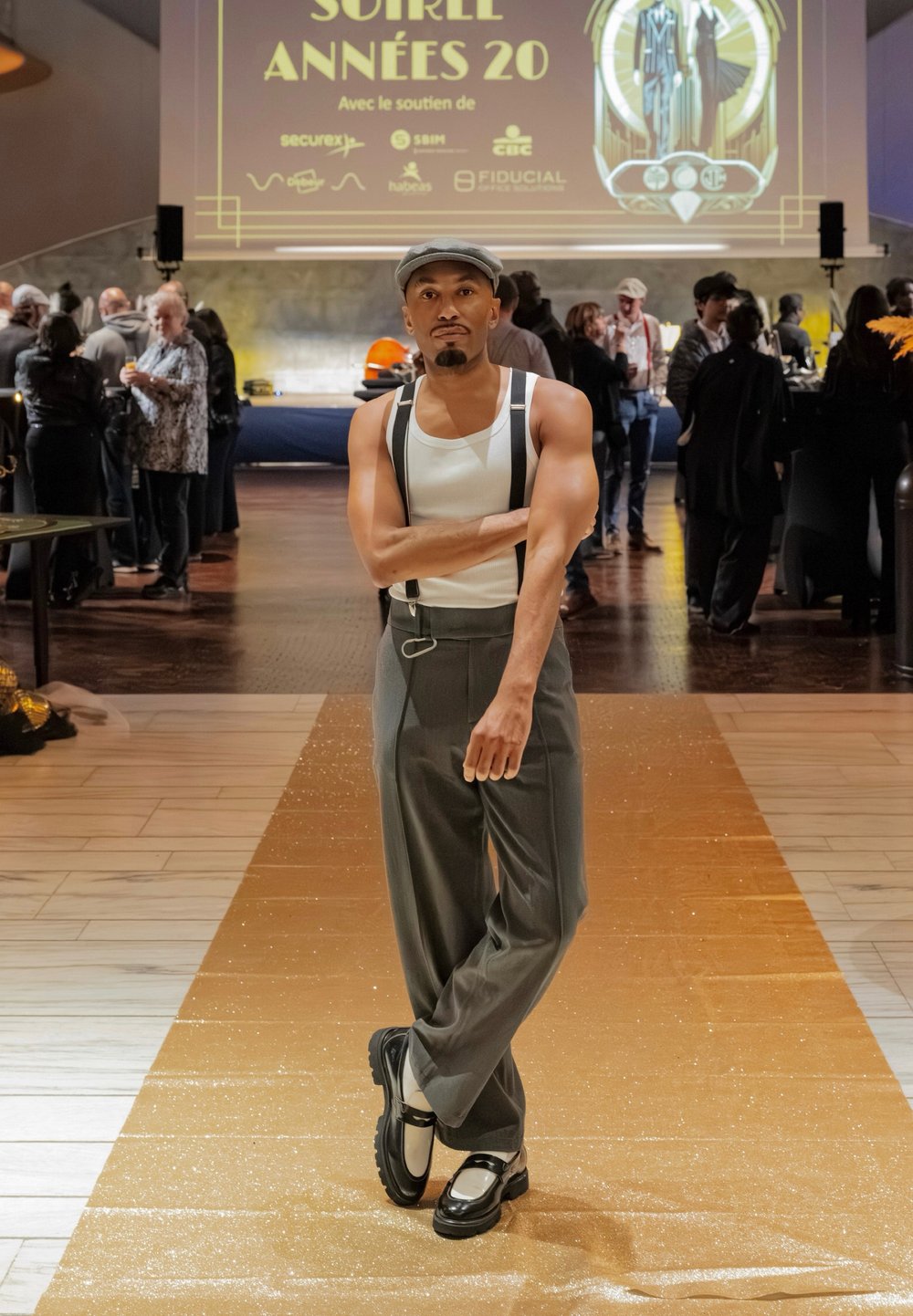 Calças cinzentas de cintura alta com suspensórios, top branco e sapatos pretos com um detalhe branco; situadas numa passarela dourada cintilante.