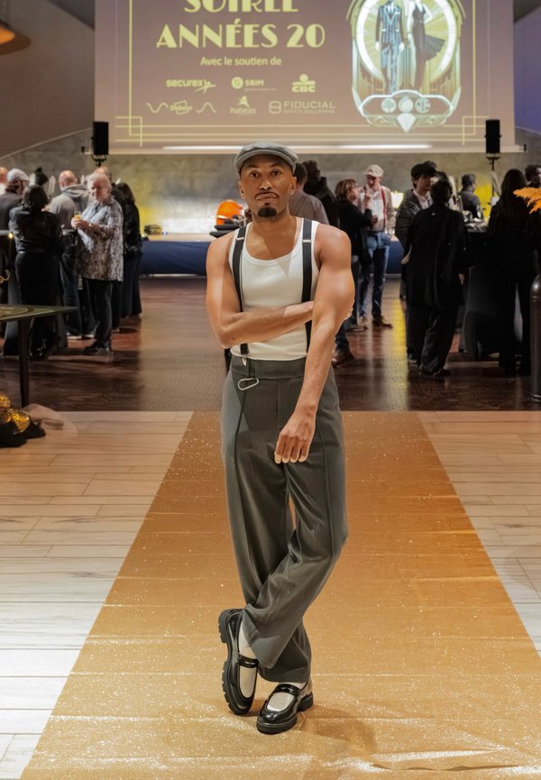 Grey high-waisted trousers with suspenders, white tank top, and black shoes featuring a white accent; set on a glittery gold runway.